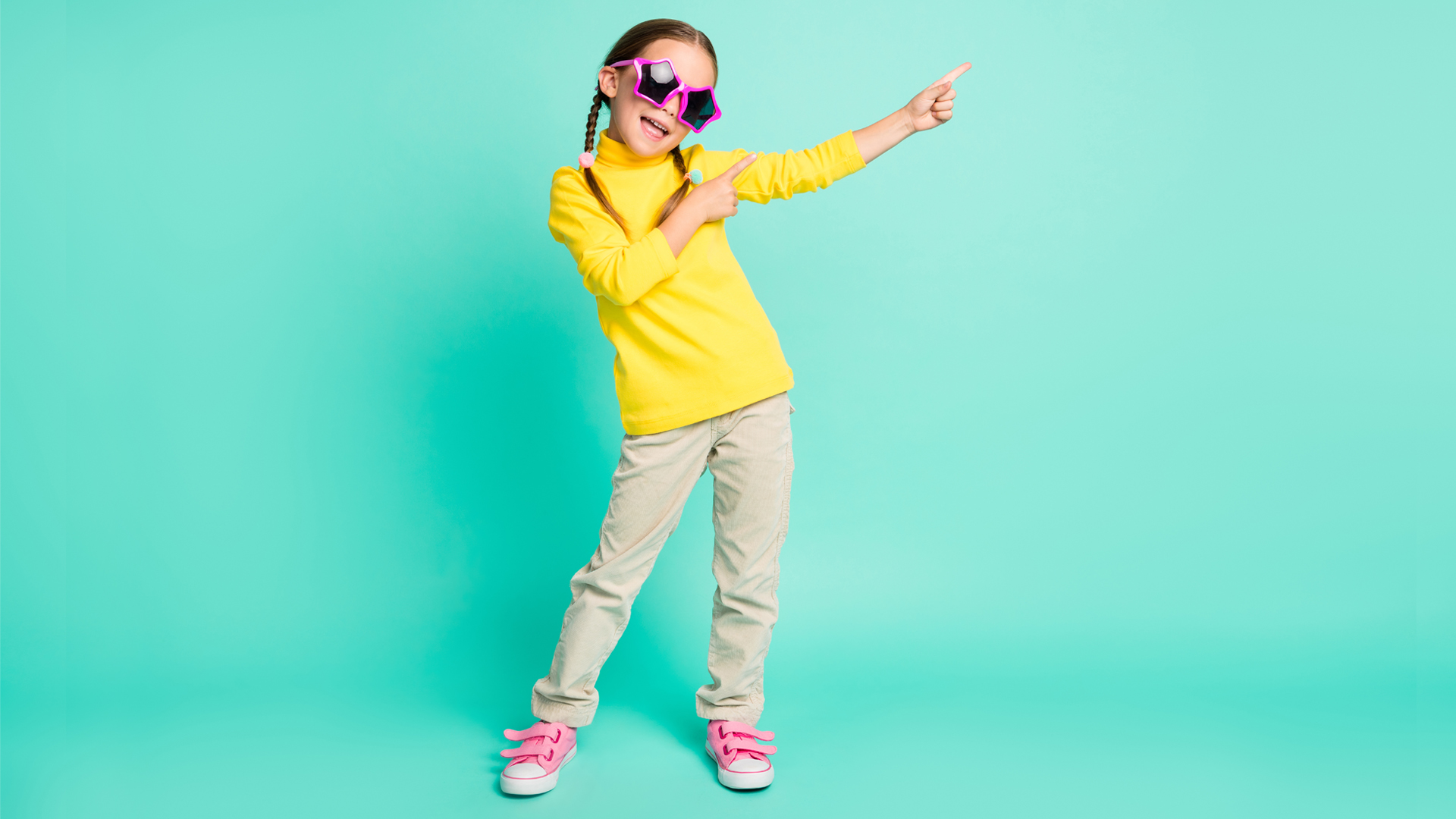 Photograph of a young girl in a bright yellow top, grey jeans and pink, star-shaped sunglasses pointing her arms and fingers to the right, against a solid, aqua background.